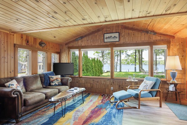 A cabin with a wooden interior, a sofa, a rocking chair, a TV, and a window overlooking a lake. - Long Lake Stone Cottage in Traverse City Frontage  Kayaks Fire pit  Pet friendly (Traverse City)