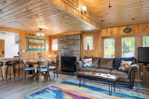 Un interior de madera con chimenea, un sofá de cuero, una mesa de comedor con sillas y una alfombra colorida.
