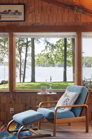 A wooden cabin with a rocking chair, a table with a glass, and a view of a lake. - Long Lake Stone Cottage in Traverse City Frontage  Kayaks Fire pit  Pet friendly (Traverse City)
