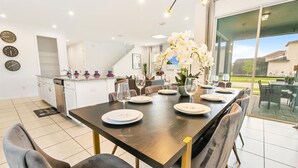 A modern dining area with a dark wooden table set for six, surrounded by grey upholstered chairs. A large window offers a view of a residential area. - Spacious 6-Bedroom Home with Private Pool & Spa in Solara Resort – Just 7 Miles (Kissimmee)