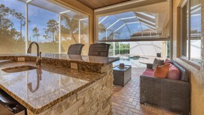 A kitchen with a granite countertop, a built-in sink, and a ceiling fan. There are wicker chairs and a cushioned bench with pillows. - Burnt Store Isles Luxury Canal Retreat - Private Pool + Spa + Outdoor Kitchen (Punta Gorda)