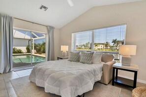 A bedroom with a large bed, two bedside tables with lamps, a view of a swimming pool, and a balcony.