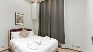 A neatly made bed with white linens and towels, a framed picture on the wall, and a dark curtain on the window. - Elegant Stay Easy Access to Heathrow & City (London)
