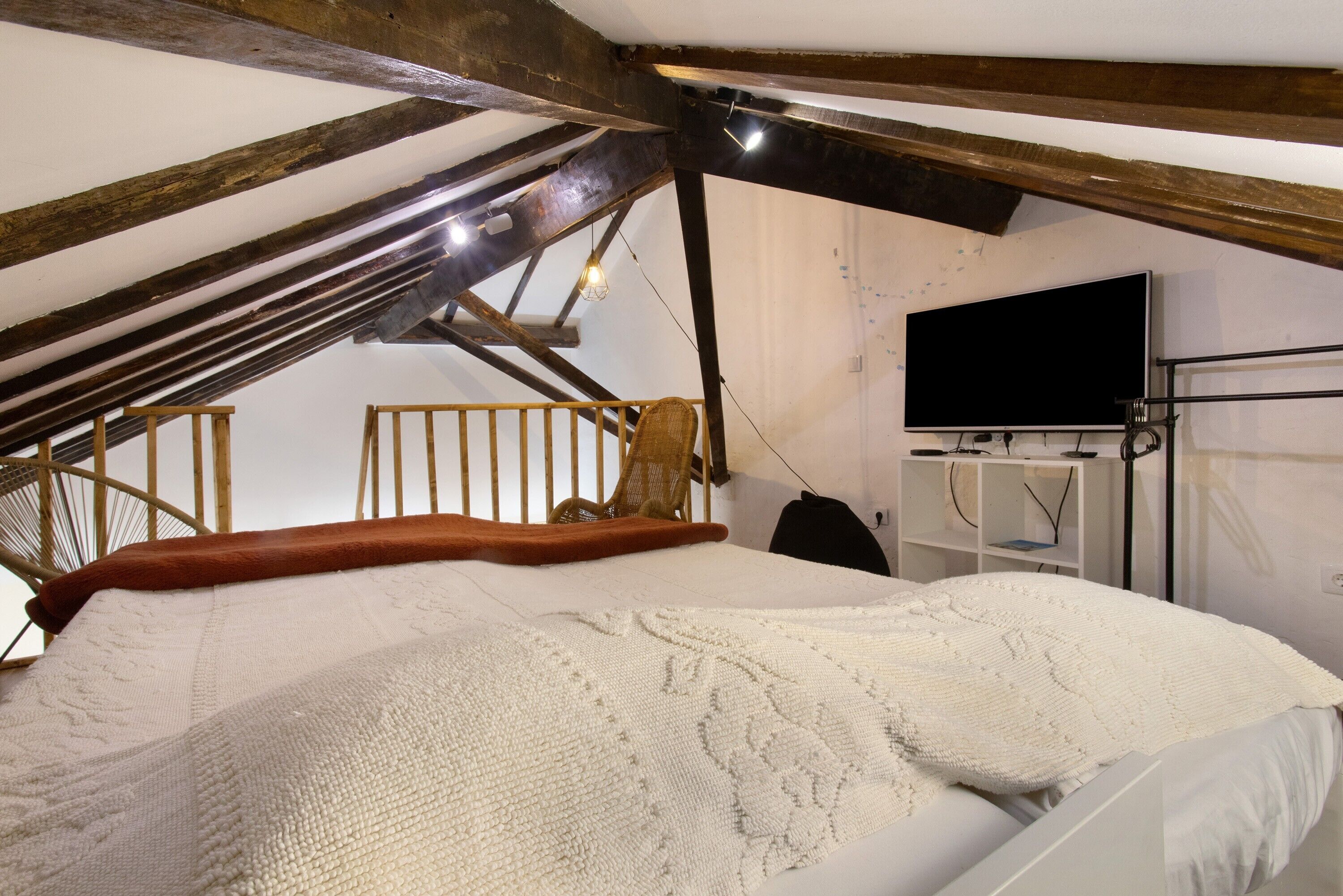 A bedroom with a sloped ceiling, a bed with a white blanket, a wooden chair, a television on the wall, and a shelf with items.