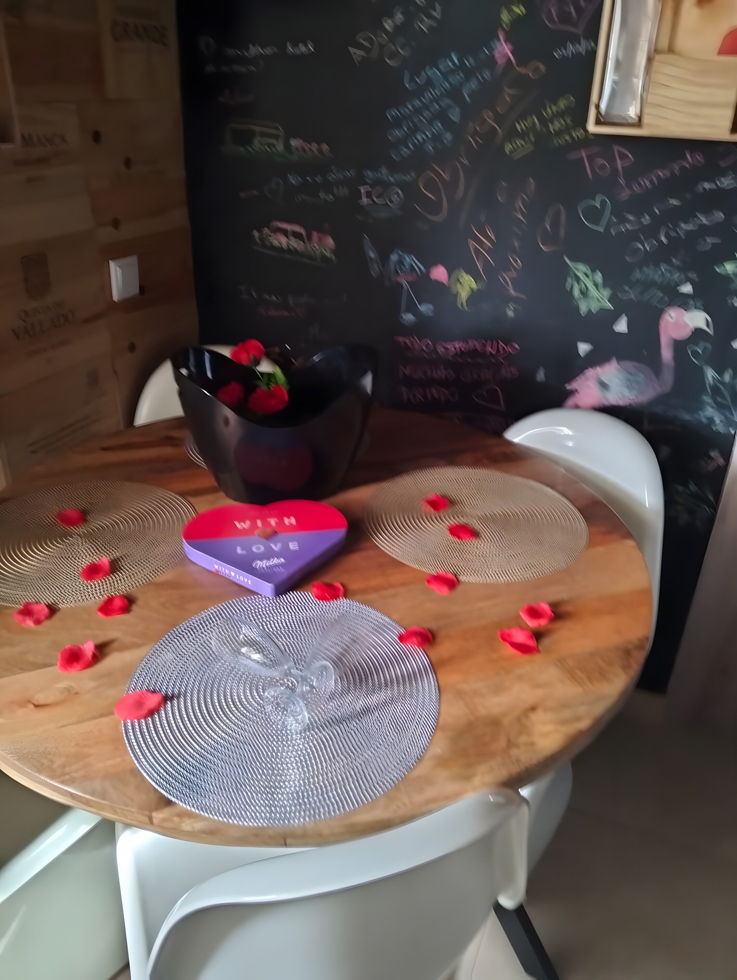 Una mesa de madera con una caja con forma de corazón, un cuenco con flores rojas y una pizarra con inscripciones.