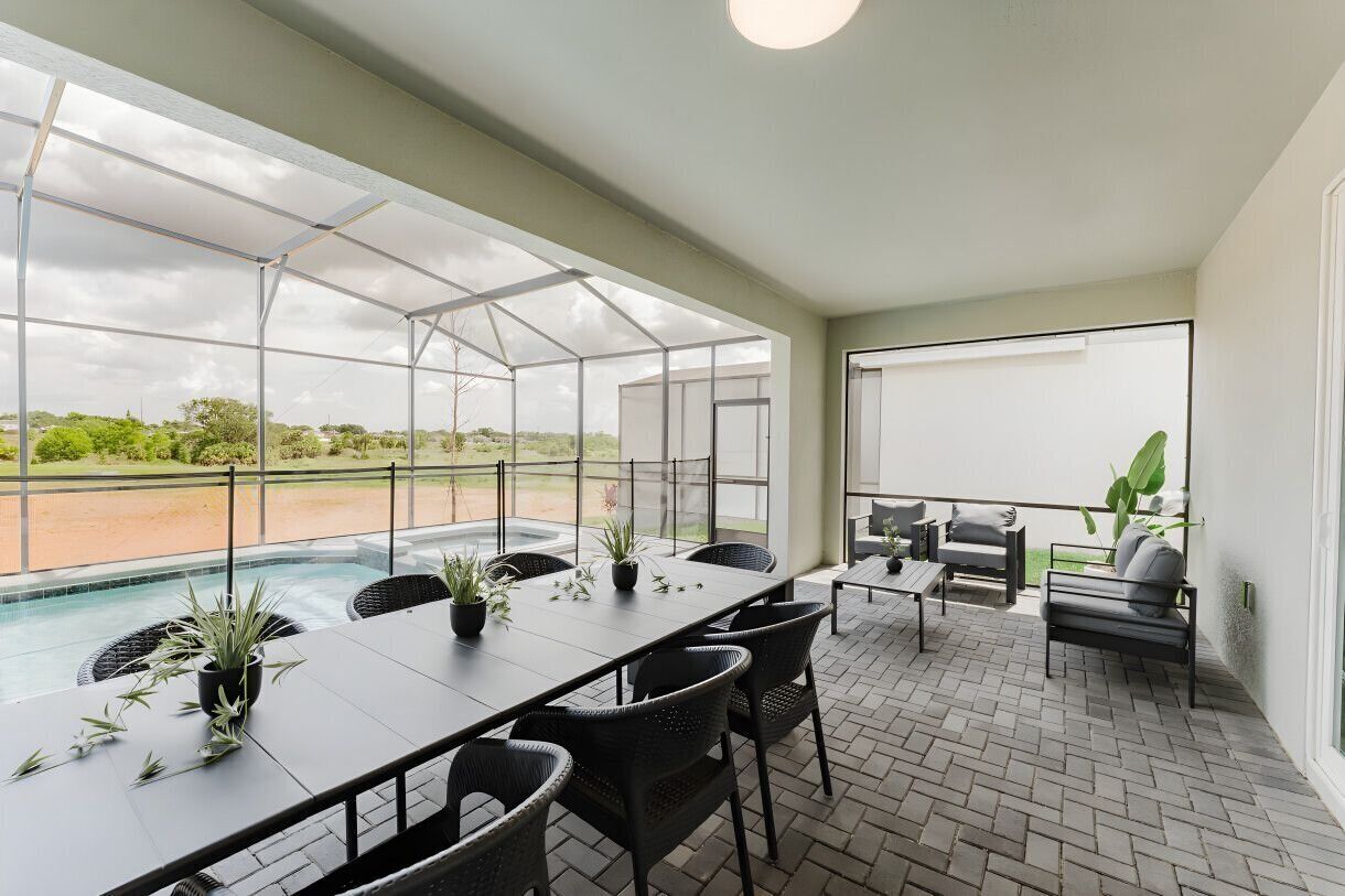 A spacious outdoor patio with a long table and chairs, surrounded by potted plants and a view of a swimming pool and greenery.