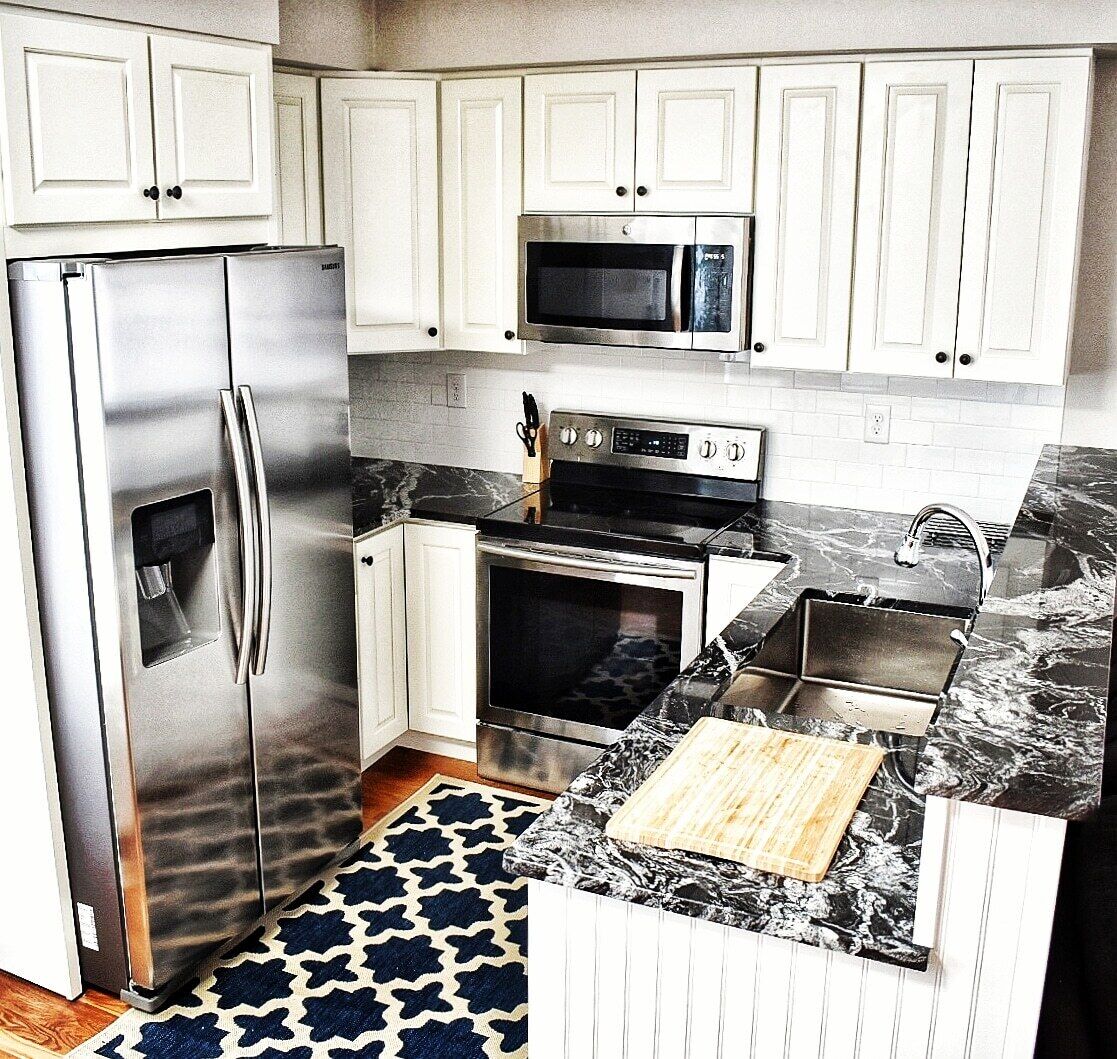 A compact kitchen with white cabinets, a stainless steel refrigerator, a microwave, an oven, and a sink with a marble countertop.