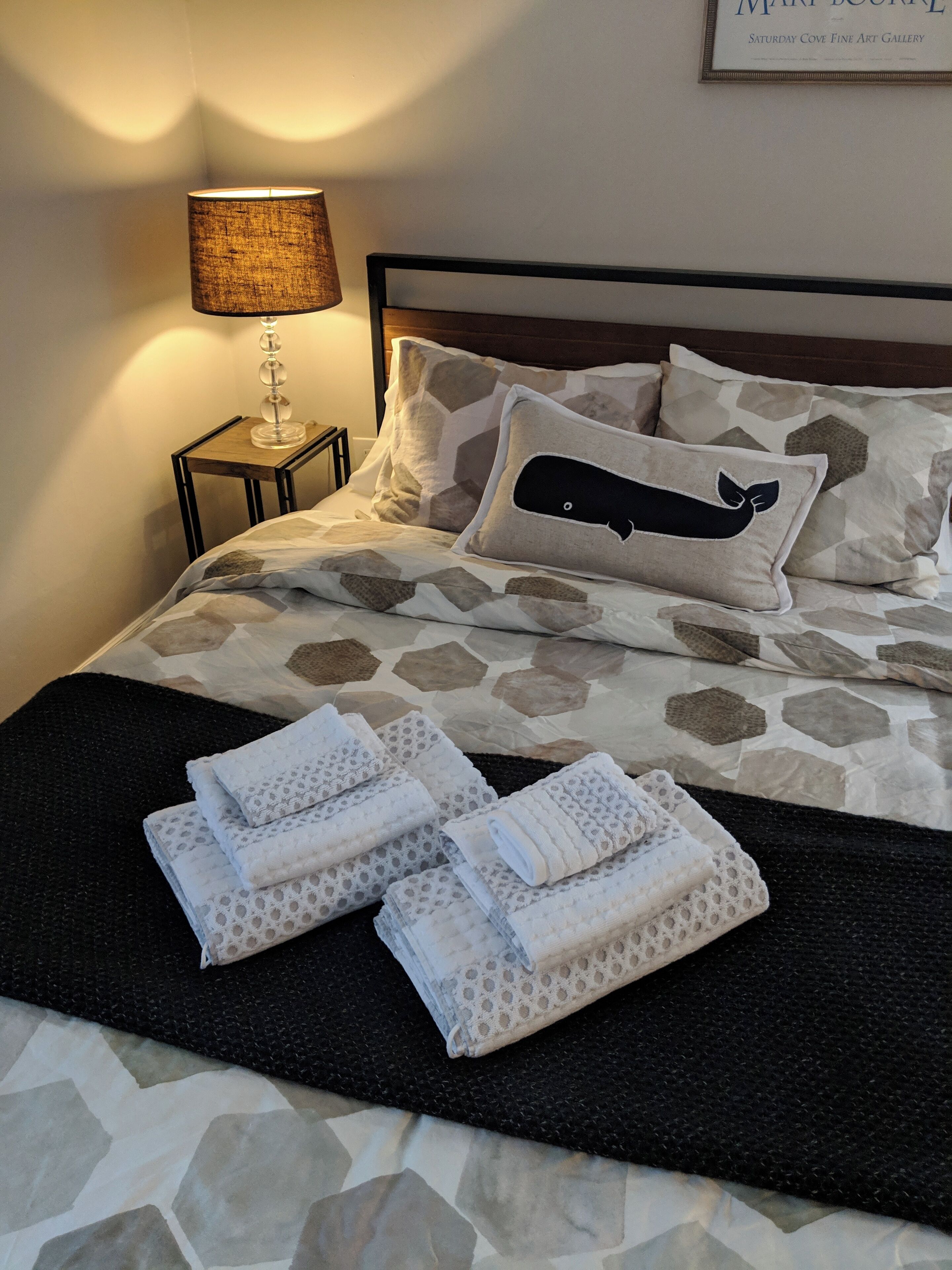 Un lit bien fait, avec une literie à motifs, un coussin en forme de baleine et des serviettes pliées sur un tapis noir.