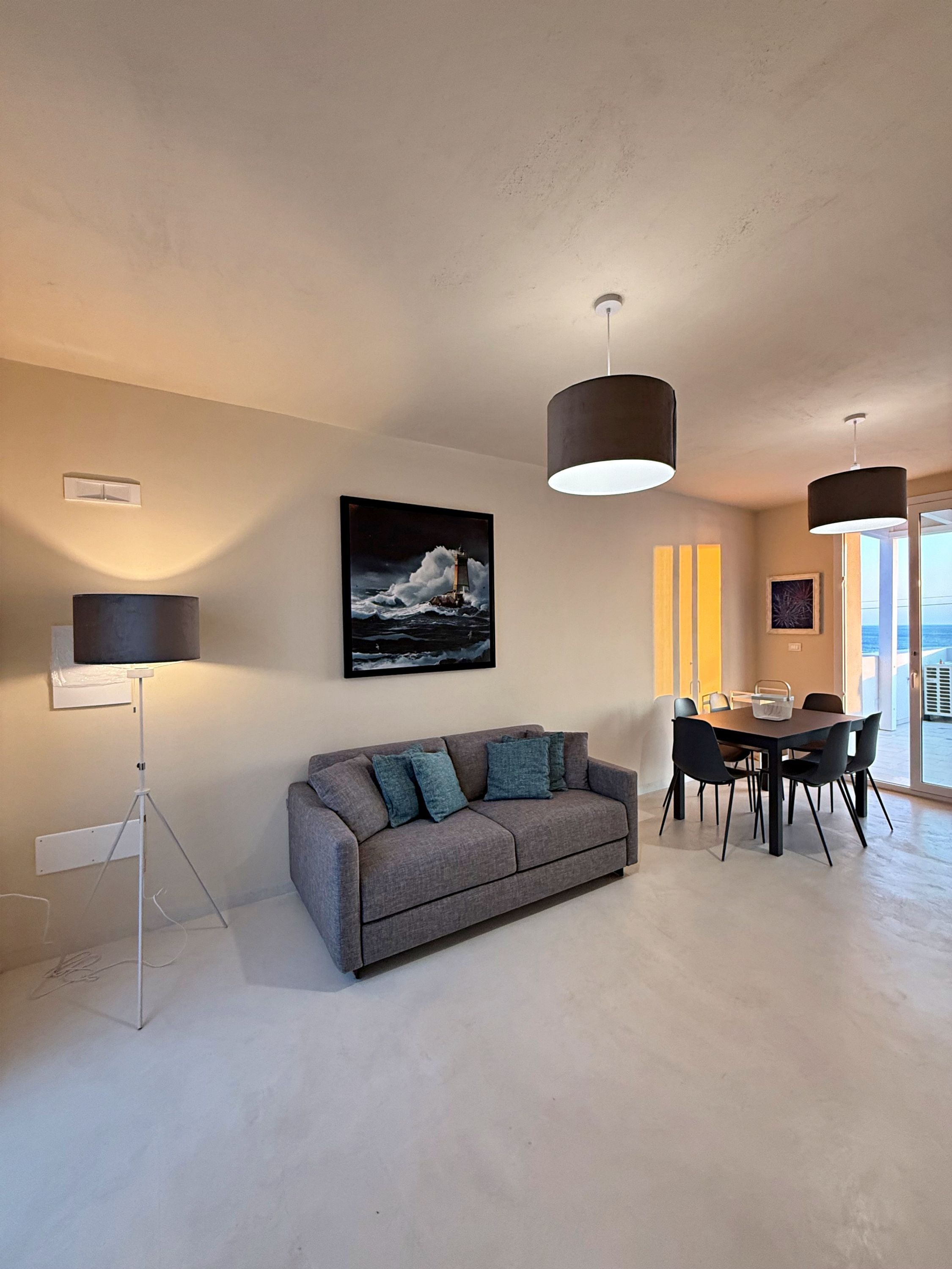 Una sala de estar moderna con un sofá gris, una mesa de comedor con sillas, lámparas colgantes y un cuadro enmarcado de un faro en la pared.