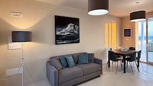 A modern living room with a gray sofa, a dining table with chairs, pendant lights, and a framed picture of a lighthouse on the wall. - Villetta Annamar - Vista Mare e Piscina Privata (Castrignano del Capo)