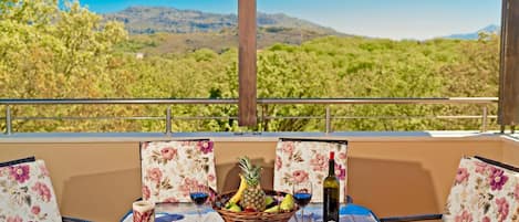 Una mesa dispuesta con una cesta de frutas, copas de vino y una botella en un balcón con sillas florales y vistas a colinas verdes.