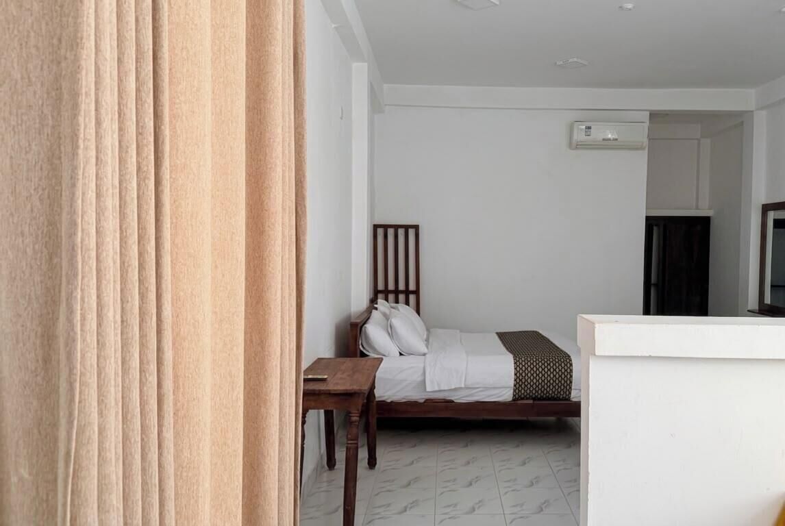 A room with a bed, a wooden chair, a small table, and a white wall with a window.