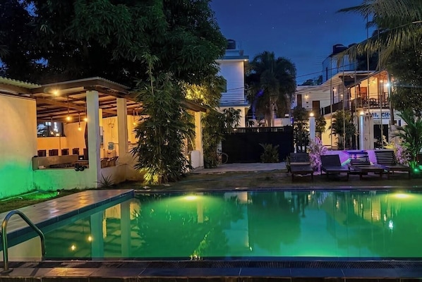 Una piscina con área techada, rodeada de árboles y edificios.
