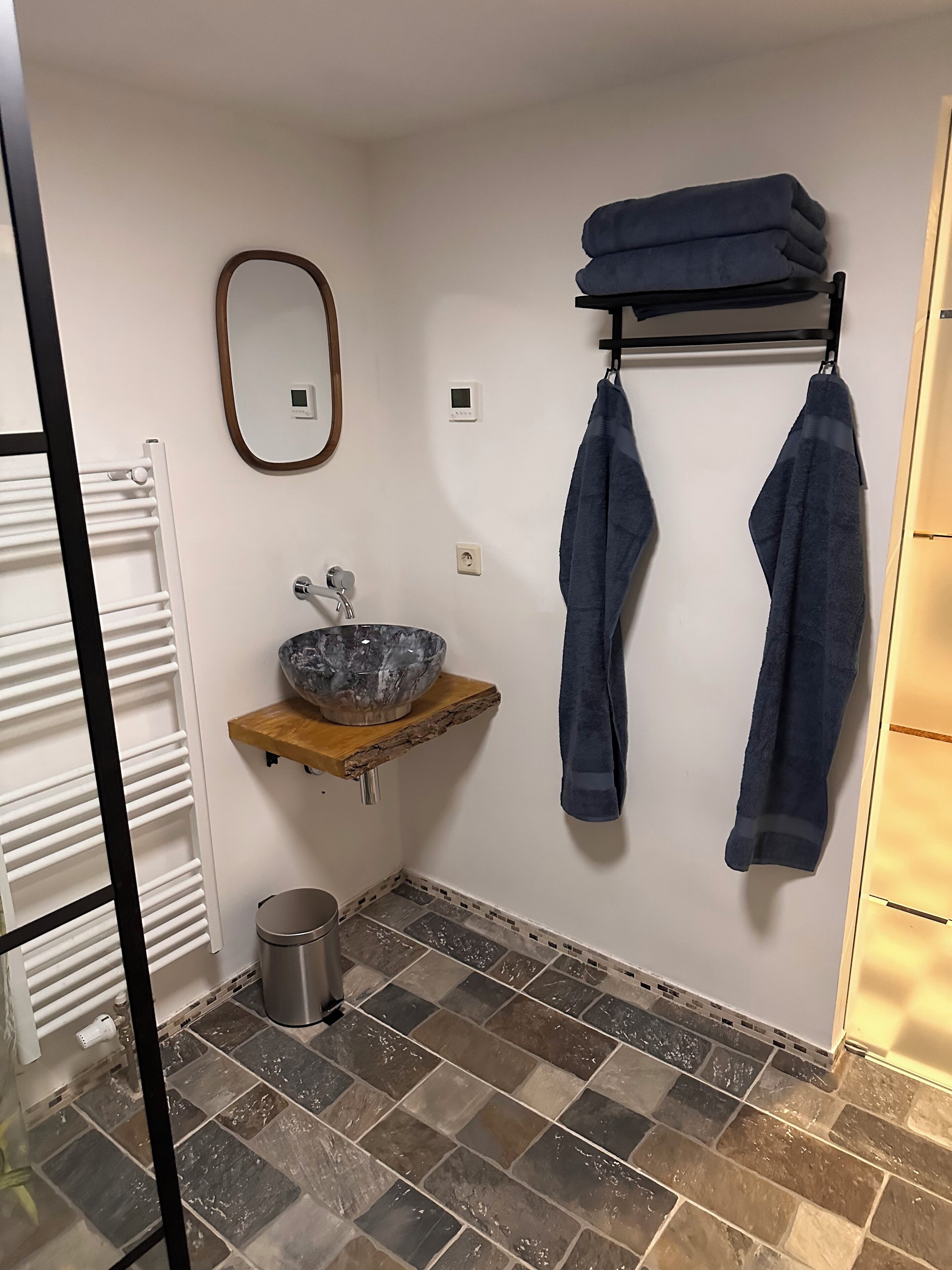 A bathroom with a marble sink, a wooden shelf, a towel rack with towels, a mirror, a radiator, and a trash bin.