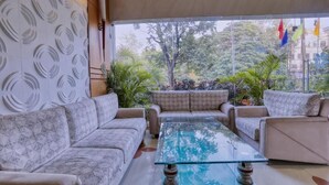 A modern lounge area with a glass coffee table, patterned sofas, and a view of greenery outside. - Hotel Amrit Residency (Indore)