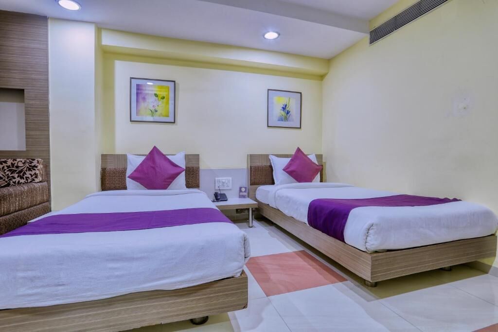 A hotel room with two beds, each with a wooden frame and white bedding. There are two nightstands with lamps, and the walls are painted light yellow.