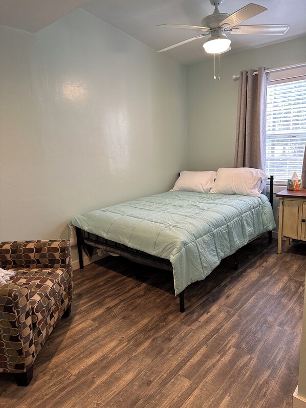 A bedroom with a bed, a wooden floor, a ceiling fan, and a window with curtains.