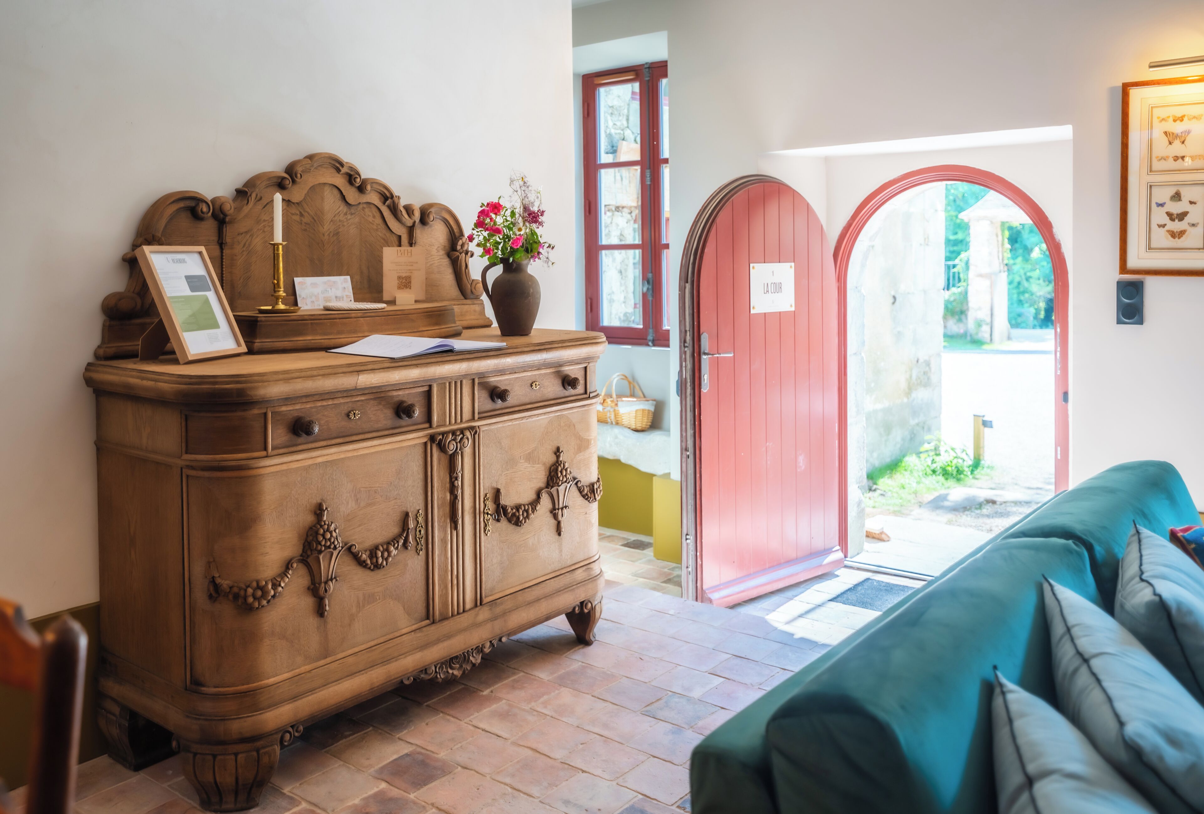Un aparador de madera con un jarrón de flores, un cuadro enmarcado y una vela. Hay una puerta roja y un sofá verde en la habitación.