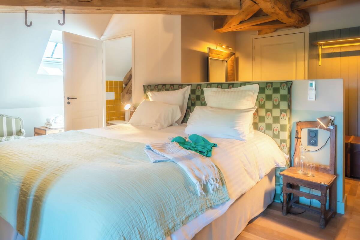 A bedroom with a large bed, wooden beams, a small chair, and a bedside table with a lamp.