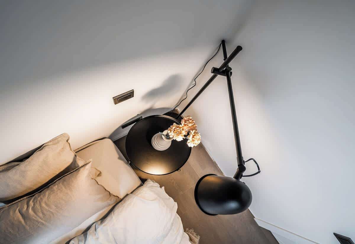 Una cama con almohadillas color beige y una lámpara negra sobre una pared blanca.