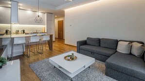 Una sala moderna con un sofá gris, una mesa de centro blanca y una cocina con armarios blancos y taburetes de bar.