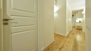 A hallway with wooden flooring and white walls, leading to a room with a window and a small table.