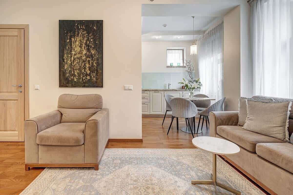 Una sala de estar con un sofá color beige, una mesa redonda pequeña y un tapete estampado. Hay una zona de comedor con una mesa y sillas, y una cocina al fondo.