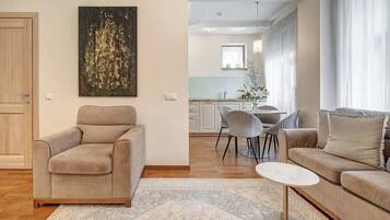 Una sala de estar con un sofá color beige, una mesa redonda pequeña y un tapete estampado. Hay una zona de comedor con una mesa y sillas, y una cocina al fondo.