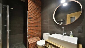 A modern bathroom with a glass shower enclosure, a white toilet, a wooden vanity with a large mirror, and dark tiled walls.