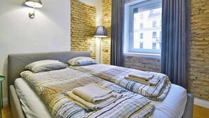 Una cama doble con ropa de cama a rayas, una lámpara de noche y vistas a un edificio a través de una ventana con cortinas.