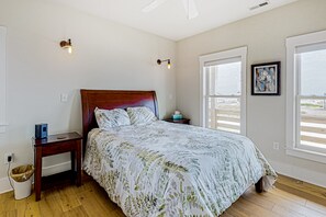 A bedroom with a wooden bed, a nightstand, a bedside table, a window with blinds, and a wall-mounted light fixture. - 3BR 4x4 ocean-view & large deck (Corolla)