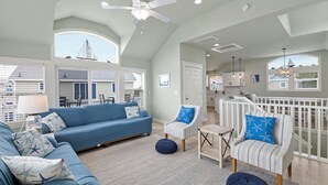 A spacious living area with a blue sofa, striped armchair, and round ottoman, a ceiling fan, and large windows. - Seaside Greetings | Hot Tub · Fenced Yard · Community Pool · Walk to Beach (Corolla)