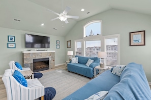 A living room with a fireplace, a television, a ceiling fan, and large windows with blinds.