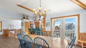 A spacious dining area with a large wooden table and blue chairs, a chandelier, and a view of the outside through large windows.