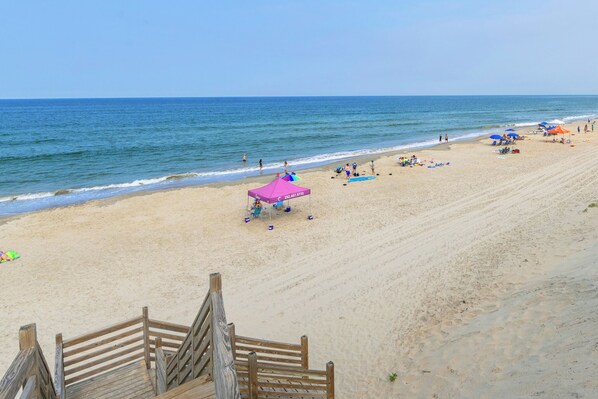 Een strand met een houten trap die naar het zand leidt, een roze parasol en mensen die ontspannen.