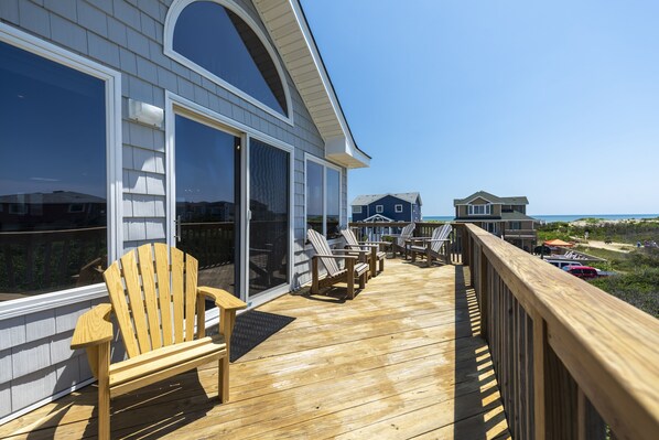 Een houten terras met stoelen en een bankje, met uitzicht op een strand en huizen.