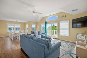 A spacious living room with a gray sofa, a dining area with white chairs, and a large TV on the wall.