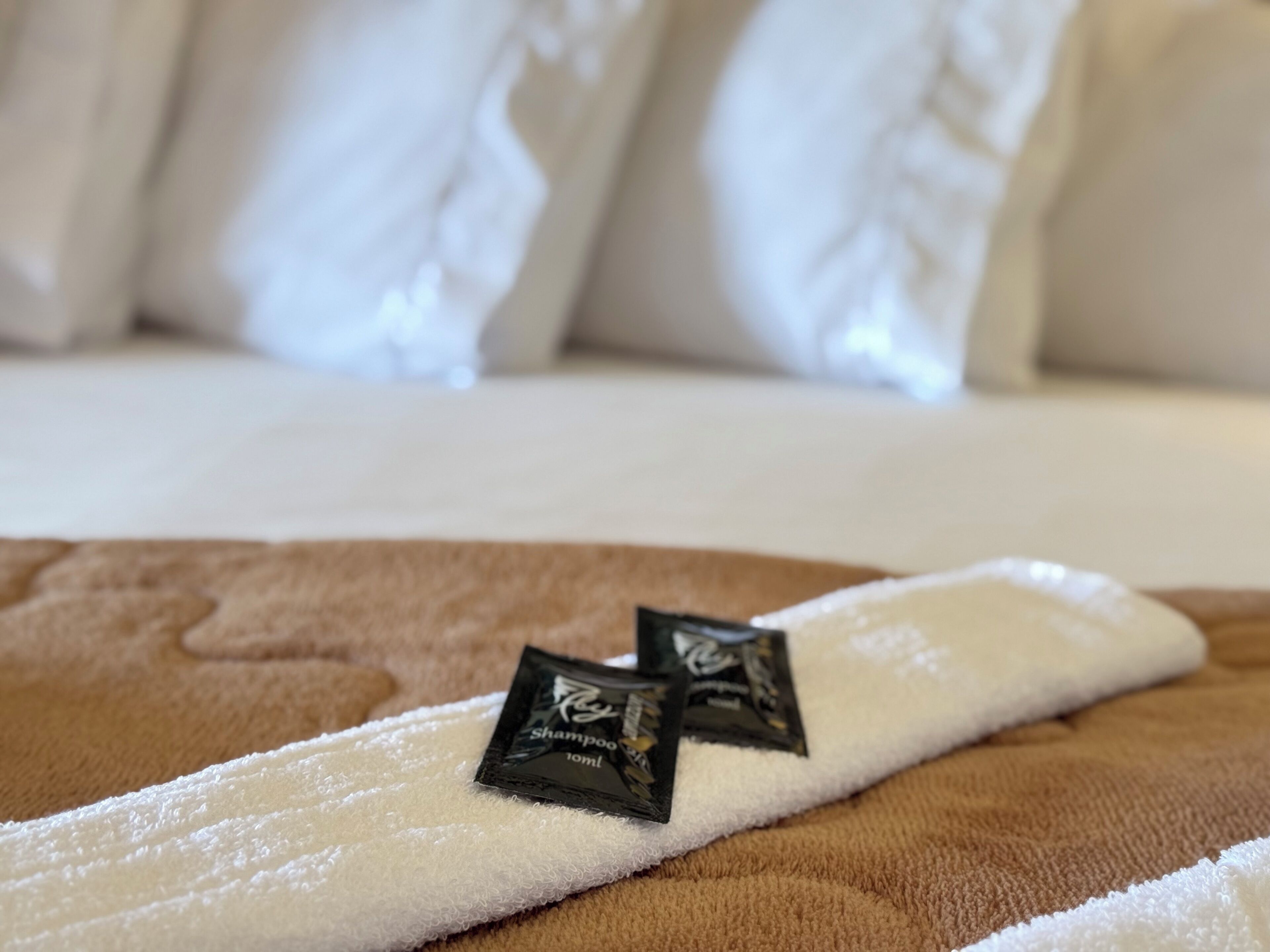 A hotel bed with white pillows and a white towel with two packets of shampoo.