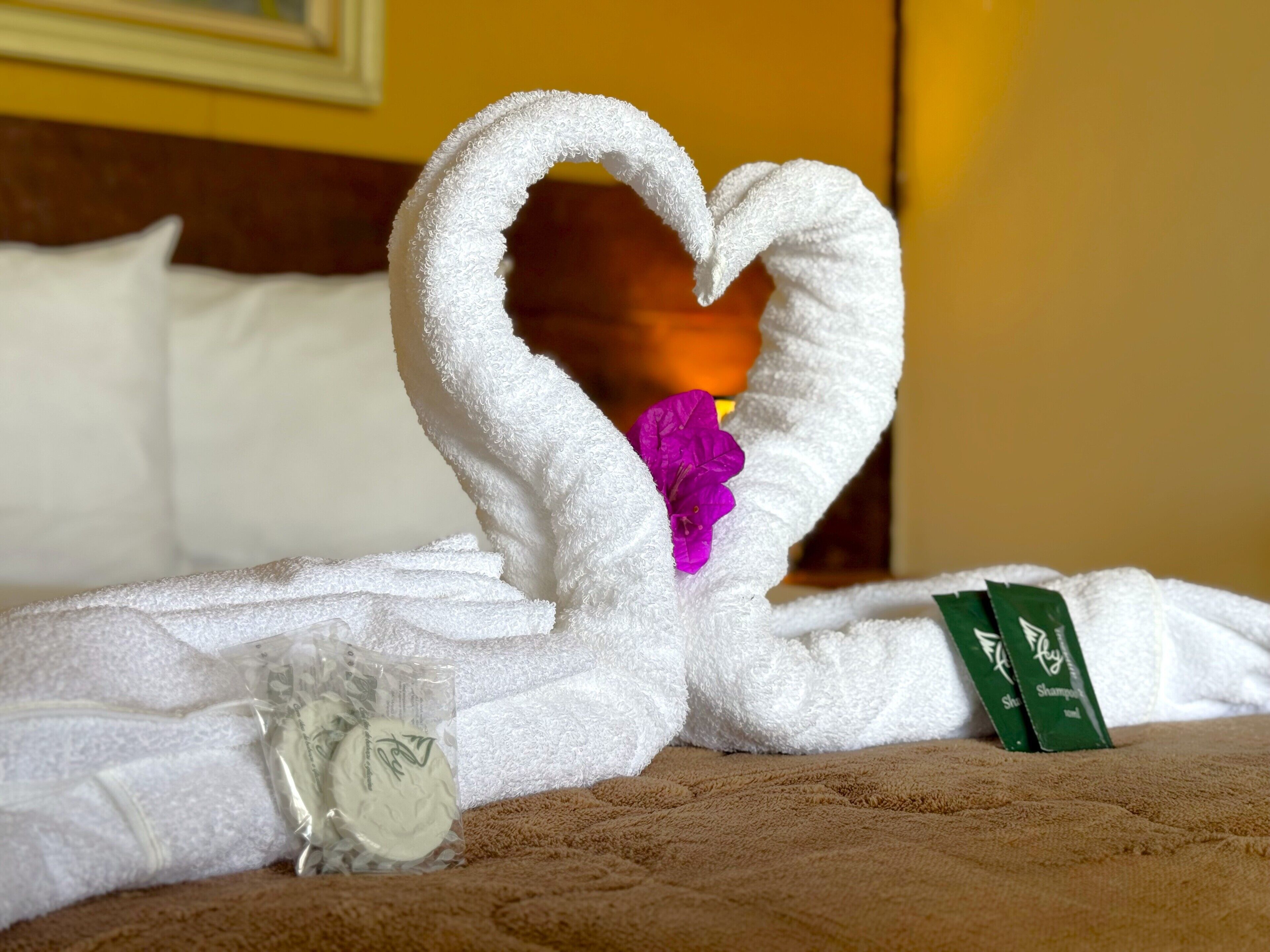 A bed with a white towel arranged in a heart shape, a purple flower, and a green bottle of shampoo.