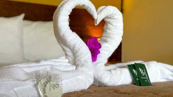 A bed with a white towel arranged in a heart shape, a purple flower, and a green bottle of shampoo.