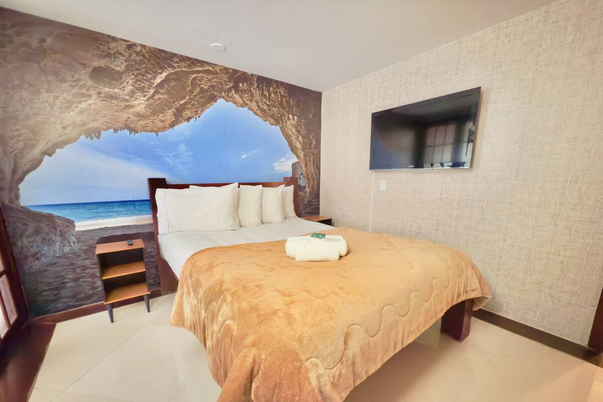 A bedroom with a large bed, a TV mounted on the wall, and a view of the beach through an archway.