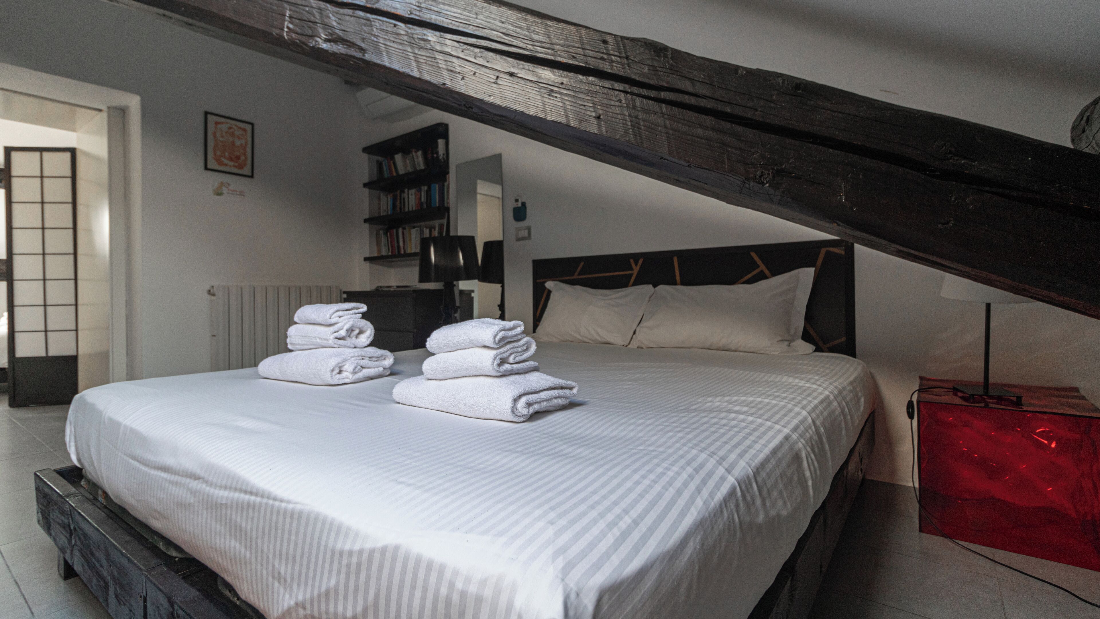 A bedroom with a large bed, a wooden ceiling beam, a bookshelf, and a red suitcase.