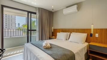 Habitación de hotel con una cama grande, cabecera de madera, mesita de noche, aire acondicionado de pared y vistas a la ciudad a través de un ventanal amplio.