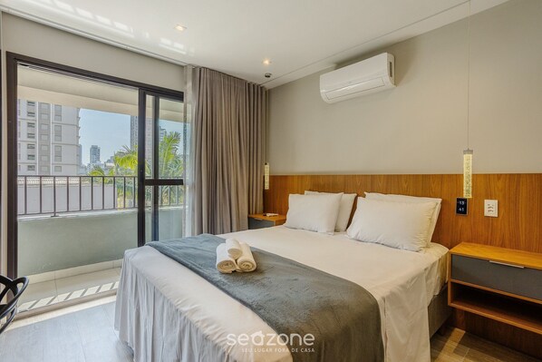 Een hotelkamer met een groot bed, een houten hoofdbord, een nachtkastje, een wandairconditioner en uitzicht op de stad door een groot raam.