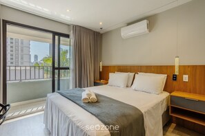 Habitación de hotel con una cama grande, cabecera de madera, mesita de noche, aire acondicionado de pared y vistas a la ciudad a través de un ventanal amplio.