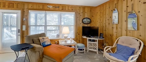 A cozy room with a wooden paneled wall, a sofa, a rocking chair, a small table with a lamp, and a television.