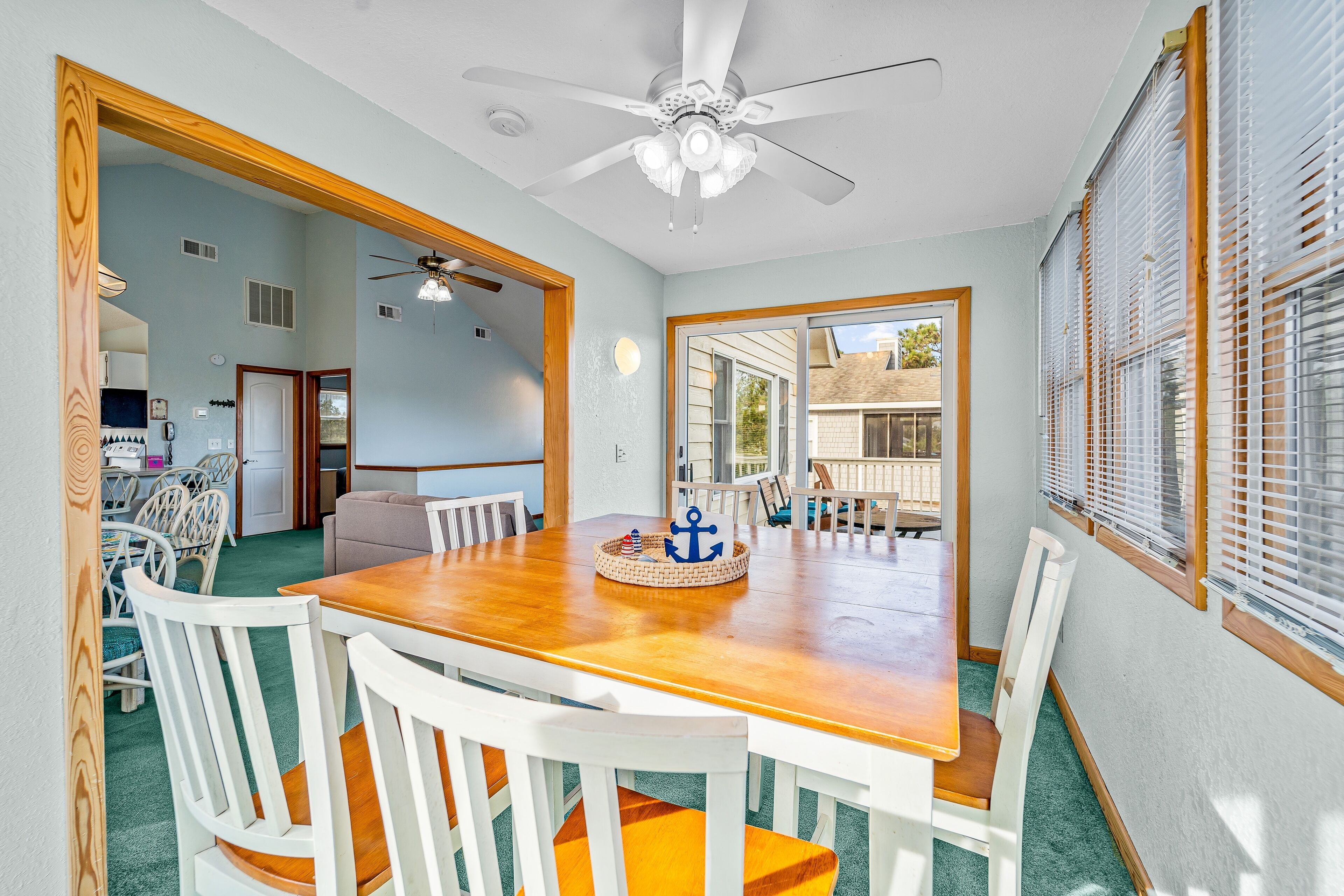 A dining area with a wooden table and white chairs, a ceiling fan, and a view through a glass door to the outside.