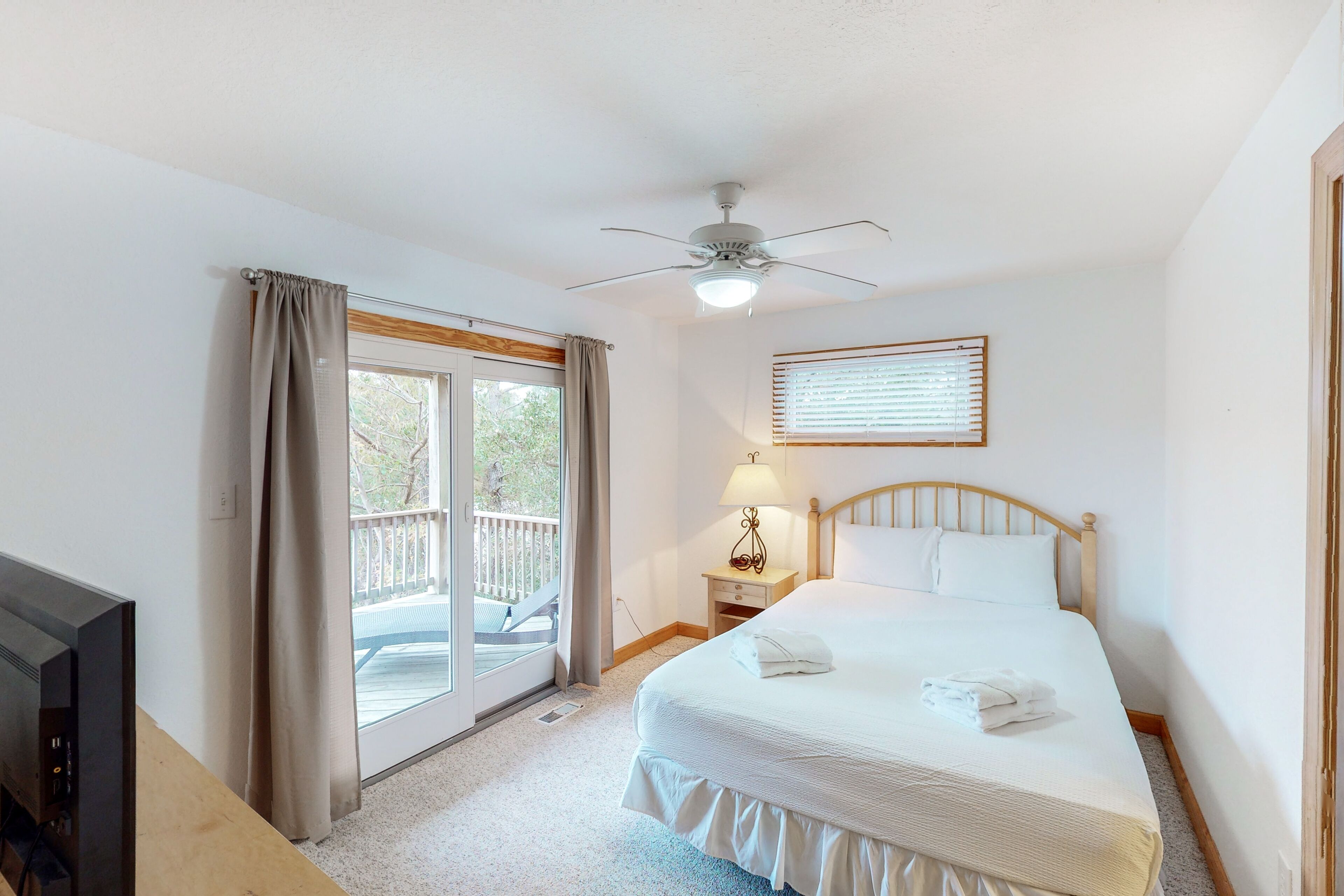 A hotel room with a bed, a TV, a ceiling fan, a window with blinds, and a balcony.