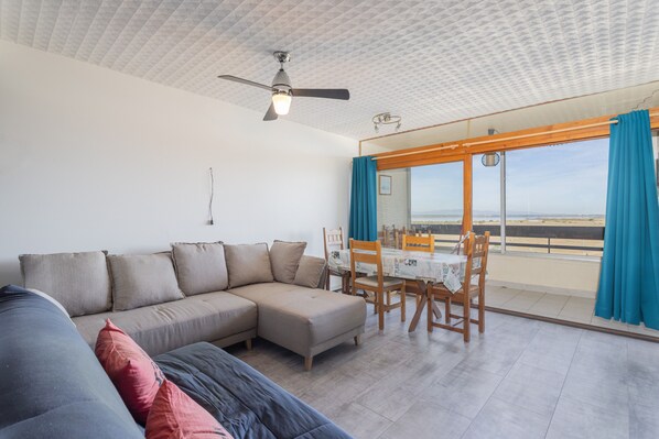 A living room with a sectional sofa, a dining table with chairs, and a balcony with blue curtains.