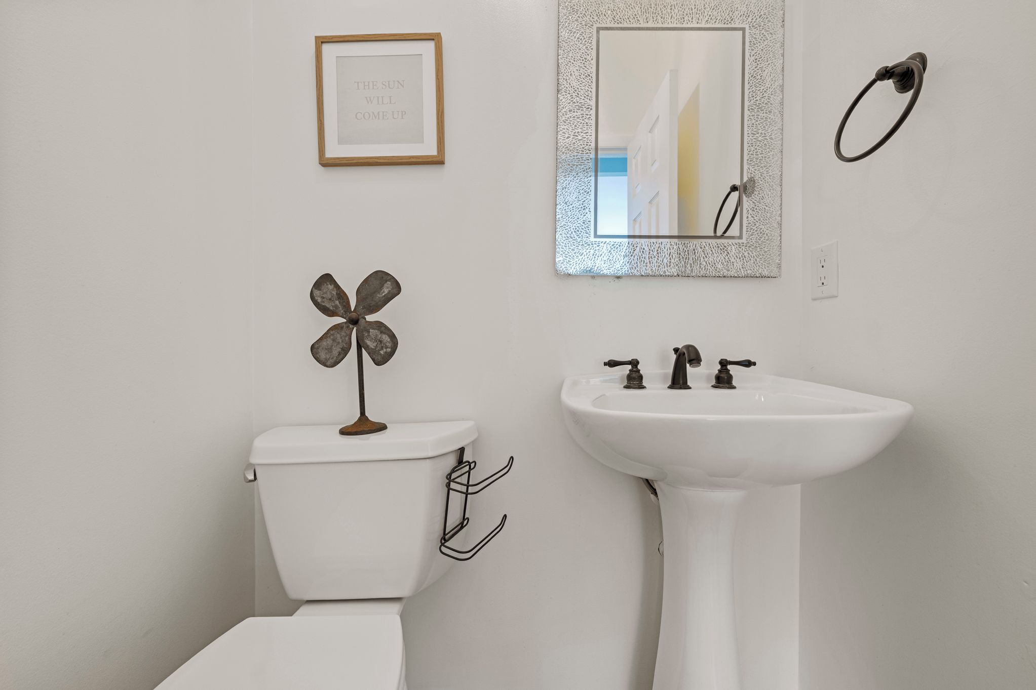Une salle de bain comprenant des toilettes, un lavabo, une fleur décorative en métal, une citation inspirante encadrée et un miroir avec un cadre texturé.